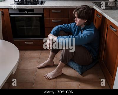 Junger Mann, der allein auf dem Küchenboden sitzt und wegschaut. Konzept von Depression, Einsamkeit, psychischen Gesundheitskämpfen und emotionalem Stress zu Hause. Stockfoto
