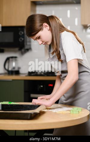 Das Kind bildet Kekse vorsichtig aus Mehl auf der Küchenoberfläche und schneidet Kekse mit dem Ausstecher in der Küche aus. Das Mädchen kocht zu Hause Stockfoto