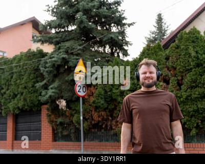 Junger bärtiger Mann in Kopfhörern, der in der Nähe einer Wohnstraße steht, mit Geschwindigkeitsbegrenzung und Grün. Entspannter Ausdruck, lässiger Lifestyle, Momentout Stockfoto