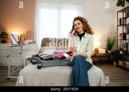 Lächelnde Frau, die am Telefon spricht, während sie den Koffer für die Reise packt Stockfoto