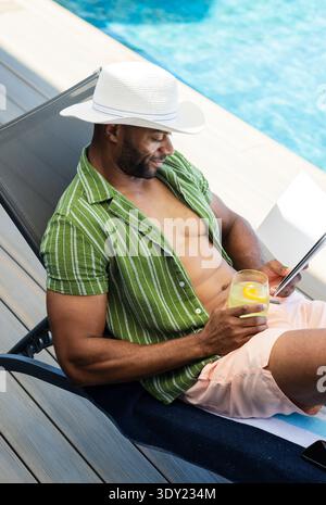 Reifer afroamerikanischer Mann, der sich auf der Liege am Pool liegend und mit einem Hut gestreiften Oberteil mit Getränk und Tablette trug. Sonnenlicht, im Freien, Terrasse, Lounge, Lesen, Limonade, Stockfoto