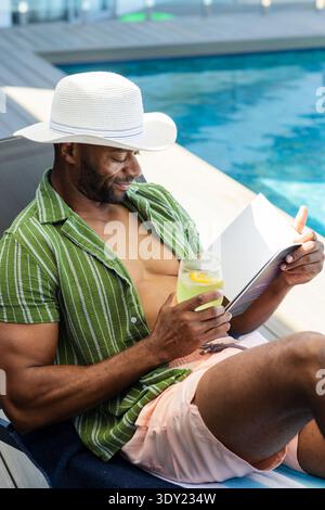 Reifer afrikanischer Mann, der am Pool liegt, einen weißen Hut trägt, ein Buch liest und ein Glas mit Zitrone hält. Pool, Freizeit, Urlaub, Sonne, gemustert, Deck, Stockfoto