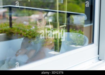 Erwachsene Frau sitzt und blickt in freistehender Badewanne durch Glasfenster mit Schaum- und Chromhahn. Badezimmer, Balkon, Grün, Reflexion, Kontempla Stockfoto