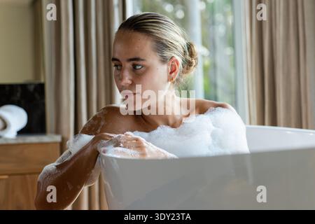Erwachsene Frau, die in einer freistehenden Wanne sitzt, mit Schaum bedeckt, Arme auf dem Rand liegend, Reifen trägt. Badezimmer, Entspannung, Ruhe, Privatsphäre, Spa, selbstpflege, Stockfoto