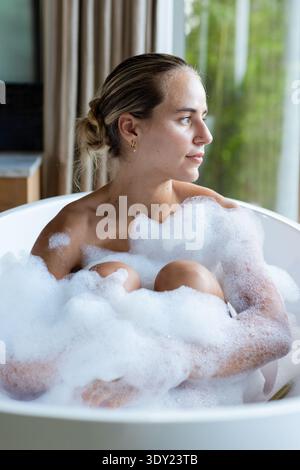Erwachsene Frau, die in einer freistehenden Badewanne mit Schaum in der Nähe des Glasfensters sitzt und goldene Körbe trägt. Bad, Blasen, Privatsphäre, Ruhe, Entspannung, natura Stockfoto