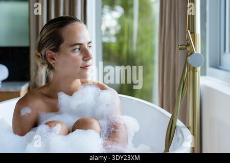 Frau, die zu Hause in einer weißen Badewanne sitzt, Knie umarmt, Schaum, Messinghahn neben dem Fenster. Freistehend, Badewanne, Vorhänge, Ruhe, Privatsphäre, Spa, Selbstpflege Stockfoto