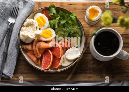Proteinreiche gesunde Frühstücksplatte mit weich gekochten Eiern, geräuchertem Lachs, frischen Gurken, Kirschtomaten, Kräutern, Zitrusscheiben und schwarzer Kaffee auf Stockfoto