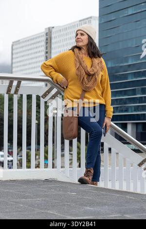 Frau mittleren Alters, die sich auf eine Metallschiene auf der Fußgängerüberführung stützt, Telefon hält, mit Senfpullover. Urban, Streetstyle, lässig, schick, Stiefel, Tasche, Moo Stockfoto