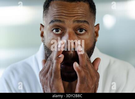 Reifer afroamerikanischer Mann, der unter die Augen drückt, mit Händen, die weißen Bademantel am Schminkspiegel tragen. Hautpflege, Gesicht, Porträt, Nahaufnahme, Selbstpflege, Mini Stockfoto