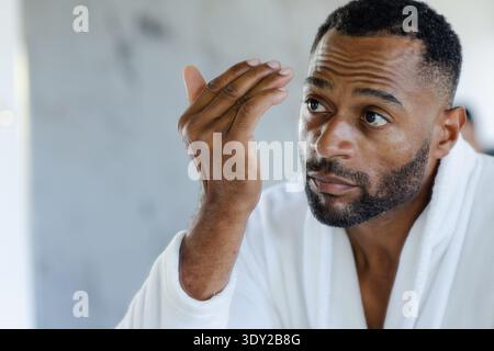 Ein afroamerikanischer Mann hebt die rechte Hand und überprüft Haare im Badezimmer mit weißem Bademantel. Porträt, reif, Bart, Gewand, Nahaufnahme, Pflege, Ruhe Stockfoto
