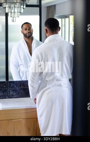 Afroamerikanischer Mann, der am Waschtisch im Bad steht und einen weißen Spiegel trägt. Kontemplativ, reflektierend, Marmor, minimalistisch, modern, Luxus Stockfoto