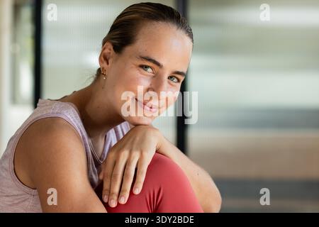 Frau sitzend, das Kinn auf dem Knie liegend, mit Tank, Leggings mit Ohrringen im Studio für Fensterrahmen. Ruhig, ruhig, weich, Kopfschuss, Innenraum, Lifestyle Stockfoto