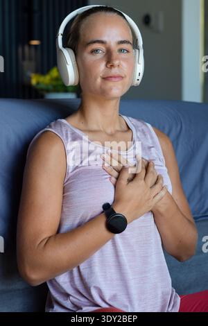 Frau, die auf einem blau-grauen Sofa sitzt, weiße Kopfhörer und rosa Tank trägt und schwarze Smartwatch zeigt. Zuhause, Lounge, Wellness, Fitness, Ruhe, minimalistisch, c Stockfoto