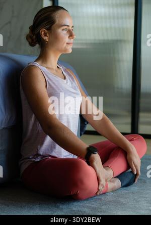 Weibliche Meditation auf Teppich neben grauem Bett, Tank und Leggings mit Smartwatch. Ruhe, Gelassenheit, Achtsamkeit, Schlafzimmer, Wellness, Zuhause, minimalistisch Stockfoto