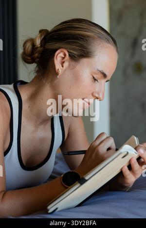 Frau, die auf dem Bett liegt, in gebundenem Notizbuch schreibt, Smartwatch und goldenen Creolen trägt. Frau, Journalarbeit, Notizen, Handschrift, Fokus, ruhig, SER Stockfoto