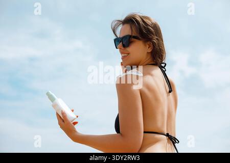 Stilvolle junge Frau in Sonnenbrille mit Sonnencreme und Sonnencreme am Strand. Konzept von Hautpflege, Sonnenschutz und gesundem Lebensstil. Weiblich Stockfoto