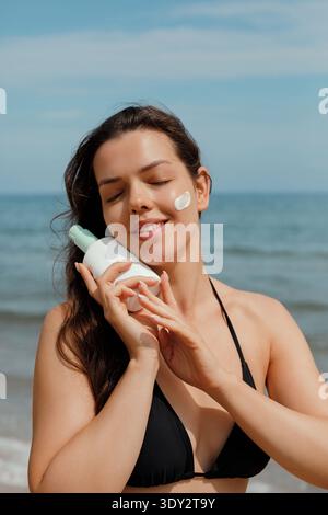 Schönheitsfrau lächelt mit geschlossenen Augen und hält Sonnencreme am Meer. Konzept von Sommer-Lifestyle, Hautpflege und Sonnenschutz. Stockfoto