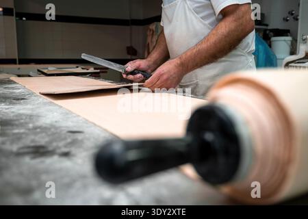 Backers Hände verwenden einen Abstreifer, um frischen Teig auf einer bemehlten Arbeitsfläche zu portionieren oder zu schneiden, wobei im Vordergrund ein Nudelstift steht Stockfoto