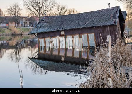 Ein altes hölzernes Bootshaus neigt sich und überschwemmt teilweise am Seeufer, seine Fenster spiegeln sich in ruhigem Wasser neben Schilf und ein ruhiges Dorf unter hellem Licht Stockfoto