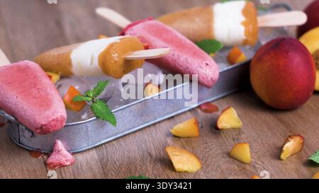 Eine erfrischende Reihe lebendiger Pfirsich- und Beerenjoghurt-Stiele liegt auf einem Eisbett in einem rustikalen Metalltablett. Natürliches Licht hebt die frostige Textur hervor Stockfoto