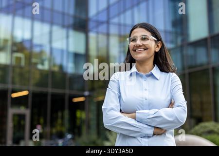 Junge Geschäftsfrau mit Brille steht selbstbewusst vor einem modernen Bürogebäude, lächelt und blickt weg, verkörpert Ehrgeiz und beruflichen Erfolg in der Unternehmenswelt Stockfoto