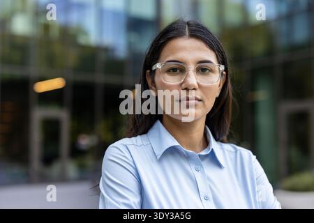 Eine junge Frau, die ein hellblaues Hemd und eine klar gerahmte Brille trägt. Selbstbewusst im Freien vor einem modernen Glasbürogebäude stehen. Darstellung von Entschlossenheit und unternehmerischer Expertise Stockfoto