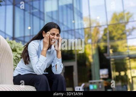 Indische Geschäftsfrau, die im Freien sitzt, ihre Schläfen unter Schmerzen durch starke Kopfschmerzen oder Migräne drückt, erschöpft und gestresst von der Arbeit, zeigt Burnout und Unbehagen Stockfoto
