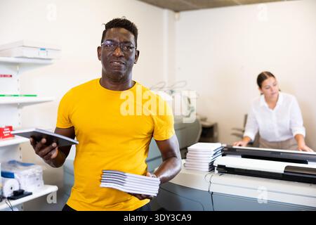 Glücklicher afroamerikanischer Typograf mittleren Alters, der neue Notizblöcke in der Mitte des Kopierzentrums zeigt Stockfoto