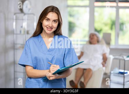 Eine Chirurg in Uniform lächelt und schreibt auf dem Klemmbrett in der Klinik mit einer Patientin, die dahinter liegt Stockfoto
