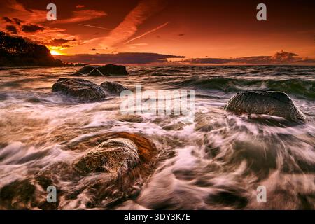 Langzeitfoto eines lebendigen, feurigen Sonnenuntergangs über einer felsigen Küste, mit glatten, milchigen Wellen, die um dunkle Felsbrocken mit einer Entfernung fließen Stockfoto