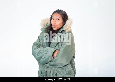 Eine fröhliche südostasiatische Frau in einem warmen olivgrünen Wintermantel mit Pelzhaube, die über weißem Hintergrund lächelt. Stockfoto