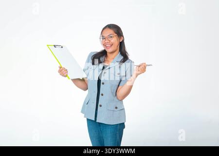 Indonesische Profi-Frau im grauen Blazer lächelnd, mit grünem Klemmbrett und Stift. In einer selbstbewussten Position auf weißem Hintergrund stehen W Stockfoto
