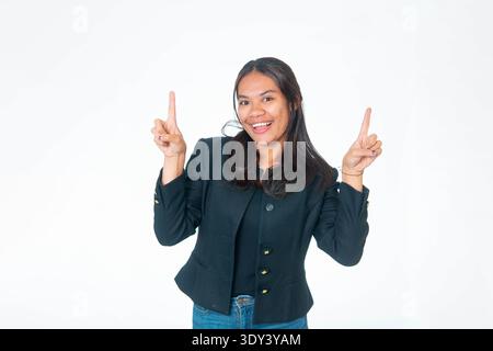 Lächelnde indonesische Frau in schwarzer Jacke, die beide Zeigefinger mit aufgeregtem Ausdruck nach oben zeigt, isoliert auf weiß. Stockfoto