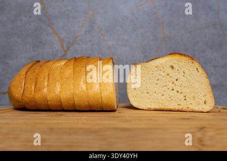 Das Brot wird auf einem Holzbrett in Scheiben geschnitten. Eine Schicht wird neben den Brotlaib gelegt. Der Hintergrund hat ein hellgraues Design mit goldenen Linien. Stockfoto