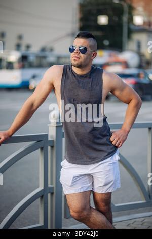 Porträt eines muskulösen Mannes, der ein Tanktop und Shorts trägt und sich an einem sonnigen Tag in einer russischen Stadt auf ein Geländer stützt Stockfoto