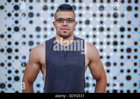 Muskulöser Sportler, der vor einer modernen Designwand steht, ein Tanktop und eine Brille trägt Stockfoto