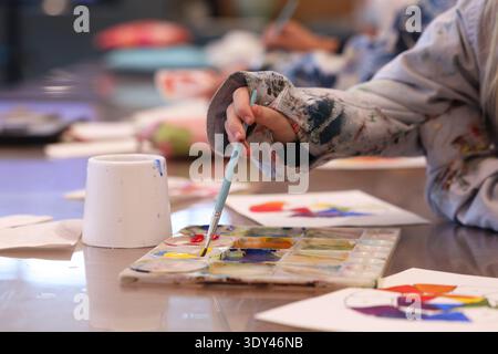 Nahaufnahme der Hand eines isolierten Kunststudenten, der einen Pinsel hält, der Acrylfarbe in einer Palette mischt. Benutzte Schürze oder Smock und authentische Kunstklasse Stockfoto