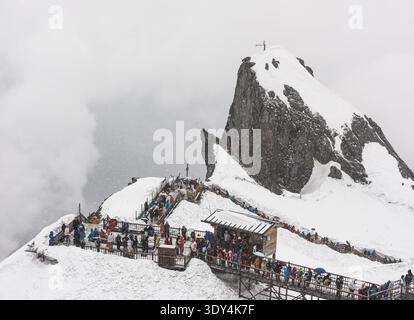LIJIANG, YUNNAN, CHINA - 12. APRIL 2025: Eine lange Reihe von Touristen in bunten, gemieteten Daunenjacken spazieren entlang einer mit Schnee A bedeckten Holzdiele Stockfoto