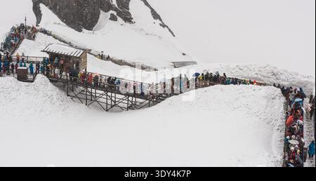 LIJIANG, YUNNAN, CHINA - 12. APRIL 2025: Eine lange Reihe von Touristen in bunten, gemieteten Daunenjacken spazieren entlang einer mit Schnee A bedeckten Holzdiele Stockfoto