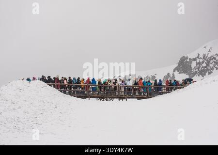 LIJIANG, YUNNAN, CHINA - 12. APRIL 2025: Eine lange Reihe von Touristen in bunten, gemieteten Daunenjacken spazieren entlang einer mit Schnee A bedeckten Holzdiele Stockfoto