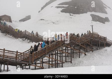 LIJIANG, YUNNAN, CHINA - 12. APRIL 2025: Eine lange Reihe von Touristen in bunten, gemieteten Daunenjacken spazieren entlang einer mit Schnee A bedeckten Holzdiele Stockfoto