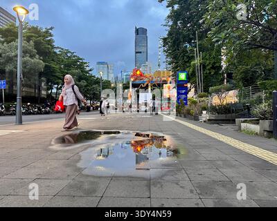 Jakarta - 5. März 2026 : Eine Frau in einem Hijab spaziert in der Abenddämmerung entlang einer belebten Straße, während Pfützen die Lichter und Wolkenkratzer der Stadt im Zentrum von Jakarta reflektieren. Stockfoto