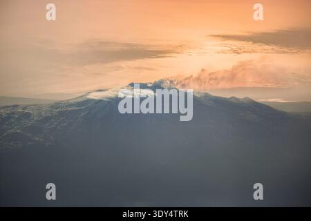 Blick aus der Vogelperspektive auf den Ätna mit einer vulkanischen Wolke, die vom Gipfel abhängt, vor einem goldenen Sonnenuntergang auf dem Ionischen Meer. Stockfoto