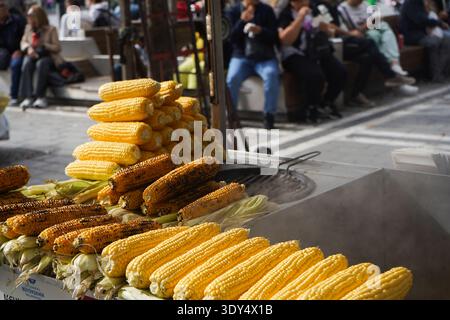 Ein lebhafter Street Food Cart in Istanbul, der frisch gegrilltes und gekochtes gelbes Mais auf dem Kolben anbietet, während die Leute im Hintergrund sitzen und sich entspannen. Stockfoto