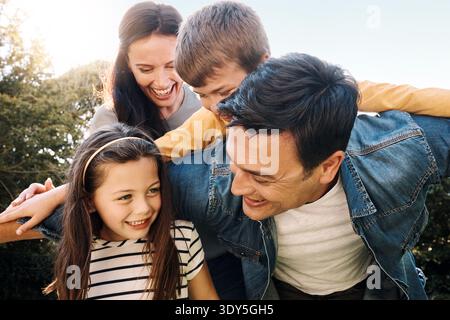Eltern, Kinder und lachen mit Huckepack im Park mit Lächeln, Binden und im Sommerurlaub im Freien. Leute, Dad und Mom mit Kindern, liebevoll oder spielerisch Stockfoto