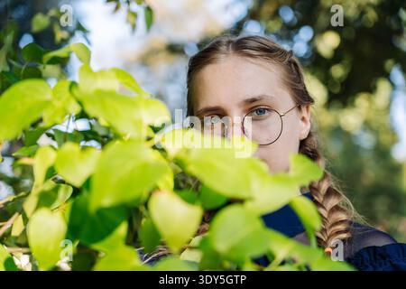 Junge Frau mit runder Brille, die draußen durch grüne Blätter schaut, blaue Augen sichtbar Stockfoto
