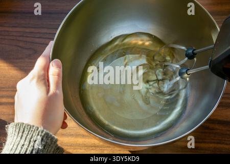 Blick von oben auf Eiweiß, das mit einem elektrischen Handmixer in einer Edelstahlschale geschlagen wird. Stockfoto
