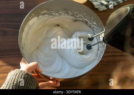 Blick von oben auf dicke glänzende Meringue, die in einer Edelstahlschale mit einem Handmixer geschlagen wurde. Stockfoto