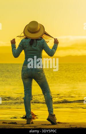 Rückansicht einer Frau in Jeansjacke und Hut am Strand zur goldenen Stunde. Sie schaut auf das ruhige Meer und hält ihre Haare. Ein ruhiger Moment der Reise, Stockfoto