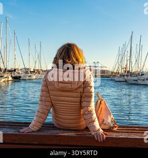 Frau, die auf einer Bank sitzt und auf die Boote im Hafen von Valencia blickt. Stockfoto